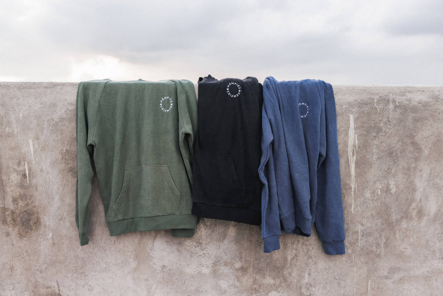 Three Sustainable Hemp and Organic Cotton Hoodies on a Wall. Olive Green, Navy Blue and Charcoal Black